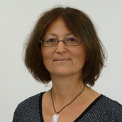 Karin Müller looks directly into the camera and smiles. She has dark brown chin-length hair and brown eyes, and wears narrow glasses and a necklace with a rectangular pendant made of white stone. She is wearing a black and white shirt with a V-neck.